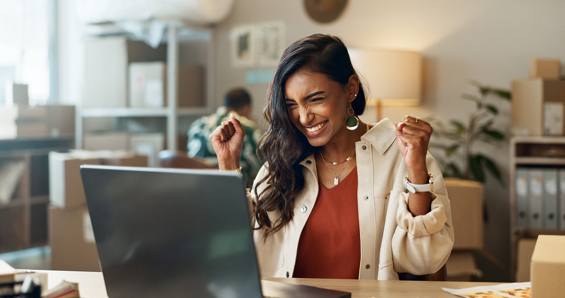 Woman sitting at desk with laptop, smiling and raising fists in celebration or success.
