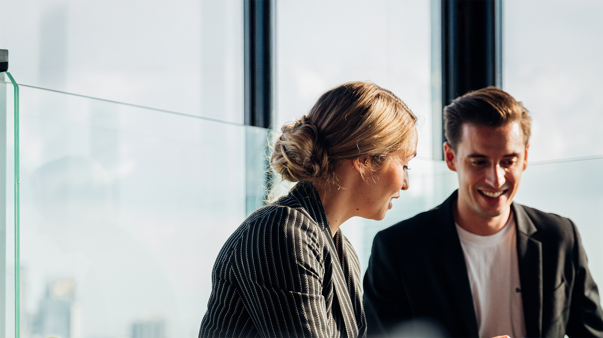 Smiling man and woman in business attire engaged in conversation near glass windows.