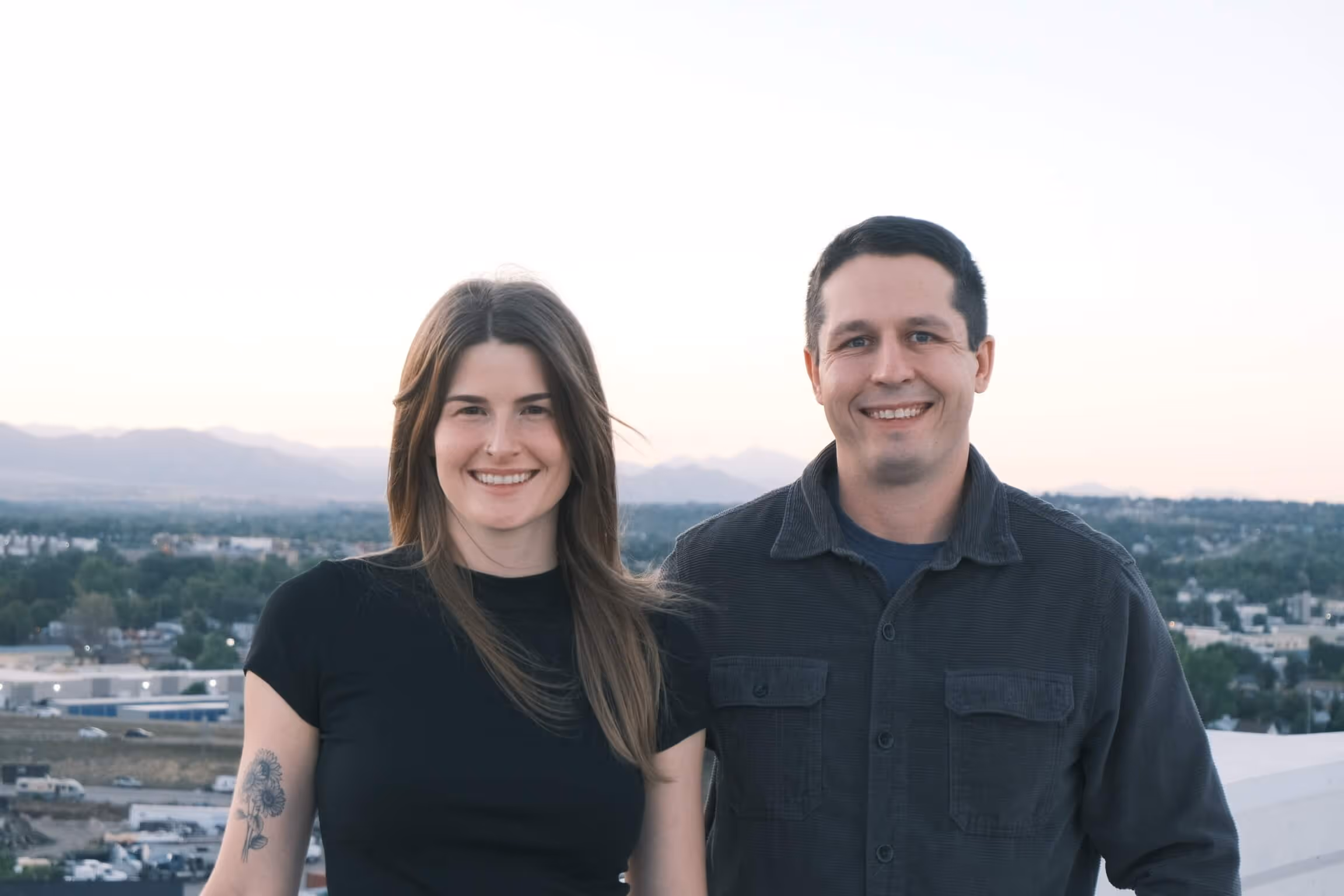 Two smiling people posing with cityscape and mountains in background