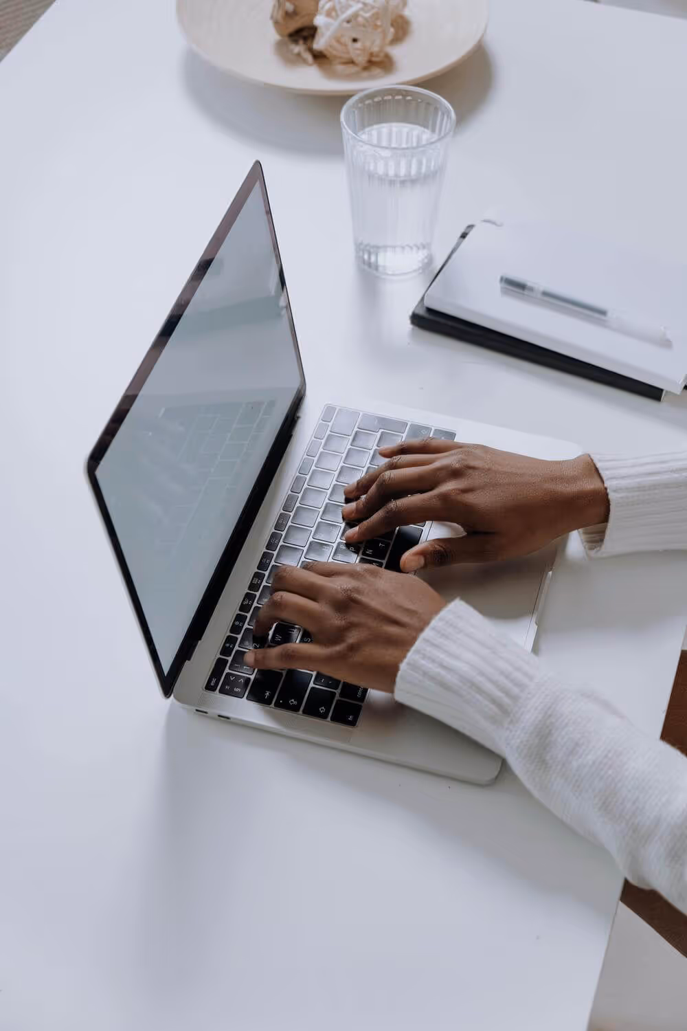 Hands typing on laptop with water glass and notebook nearby