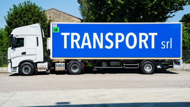 Camion bianco con rimorchio blu con la scritta bianca 'TRANSPORT srl' parcheggiato su strada asfaltata.
