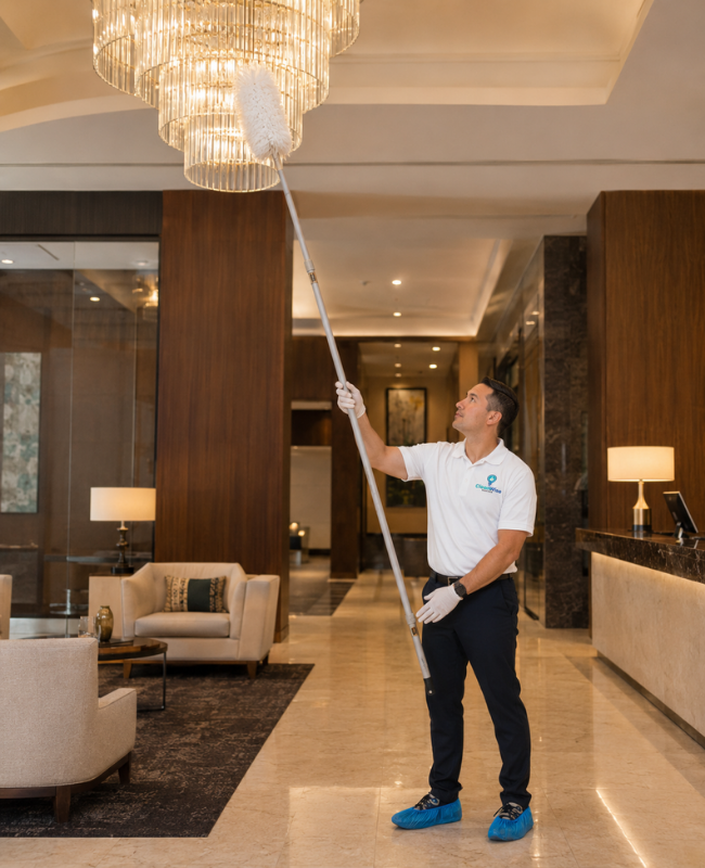 CleanWise Solutions hotel cleaning professional dusting a high chandelier in an upscale hotel lobby with a long extension duster.