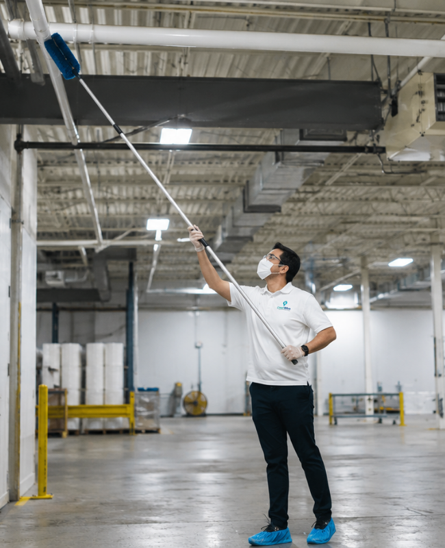 CleanWise Solutions technician using an extension duster to clean overhead pipes in a warehouse during industrial cleaning and commercial high dusting service.