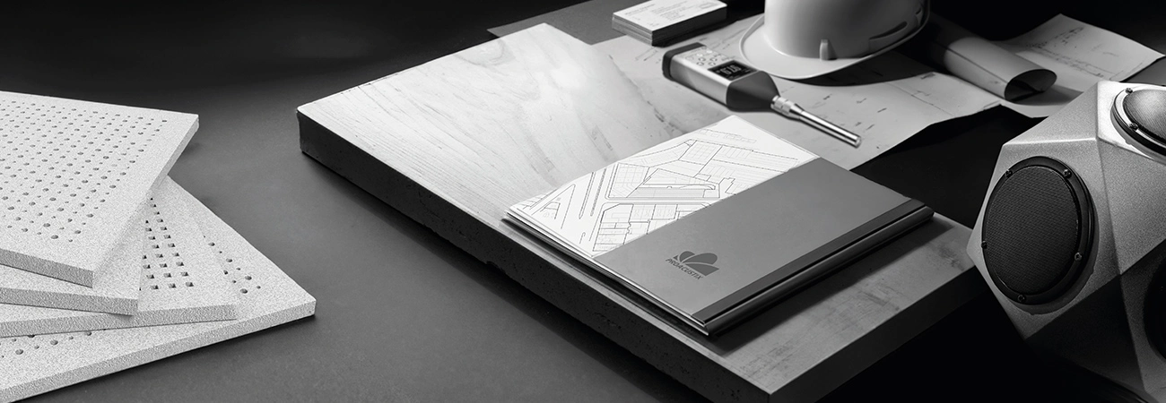 Mesa de trabajo con planos arquitectónicos, cuaderno, casco de seguridad blanco, medidor digital, muestras de materiales y bocina angular.