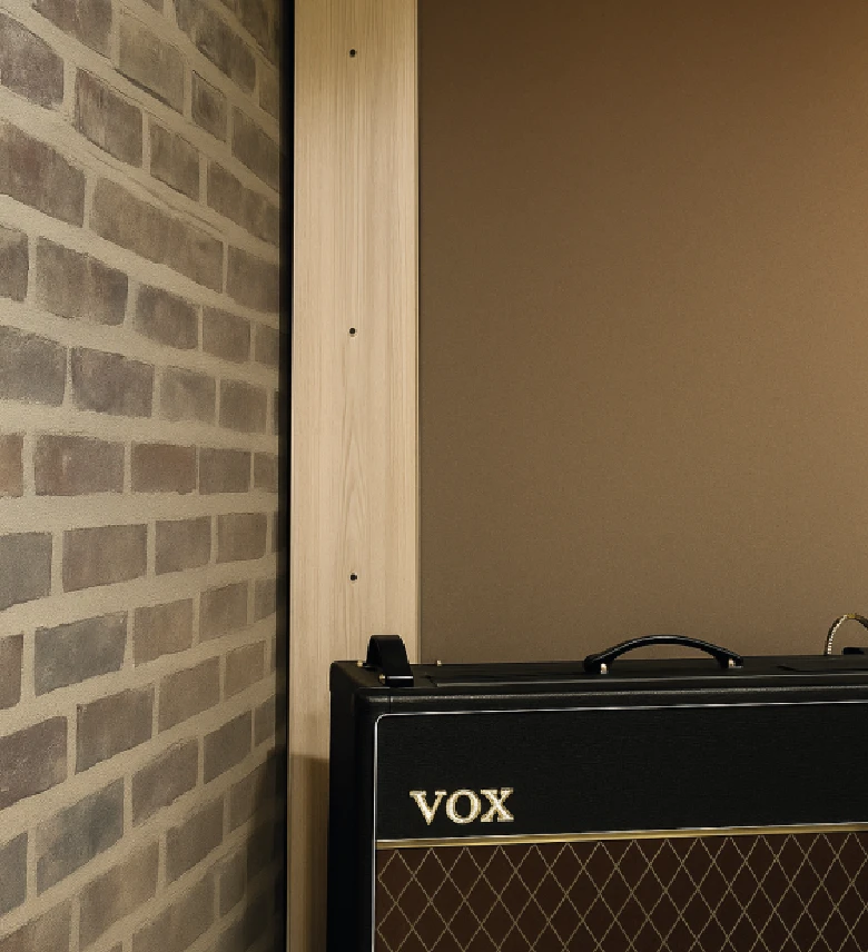 Amplificador musical VOX negro y marrón apoyado contra una pared con revestimiento de ladrillo y un panel de madera clara.