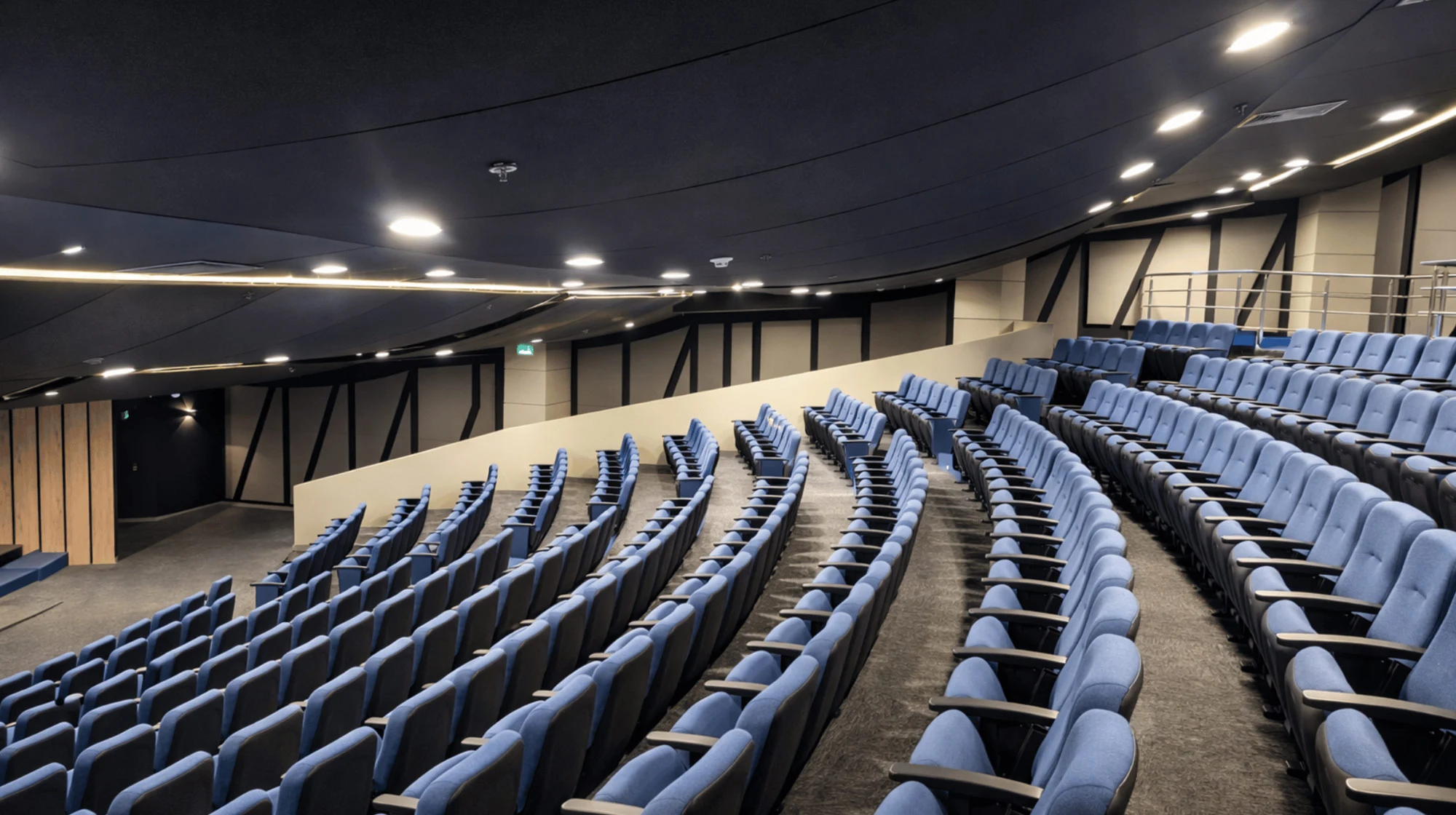 Auditorio moderno con filas de asientos azules en gradas curvas bajo techo oscuro con luces empotradas.