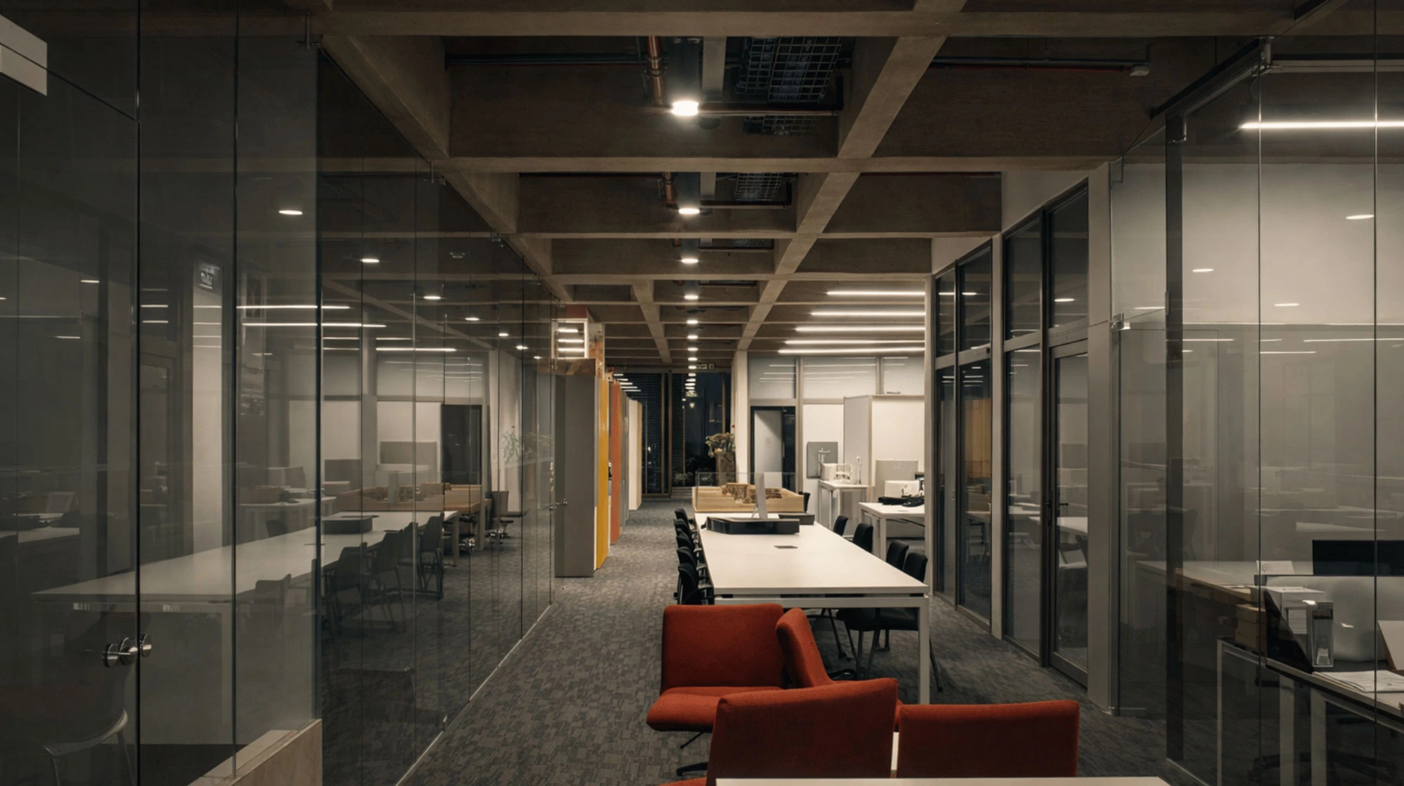 Interior de una oficina moderna con paredes de vidrio, varias mesas blancas con sillas negras y dos sillas rojas en primer plano.
