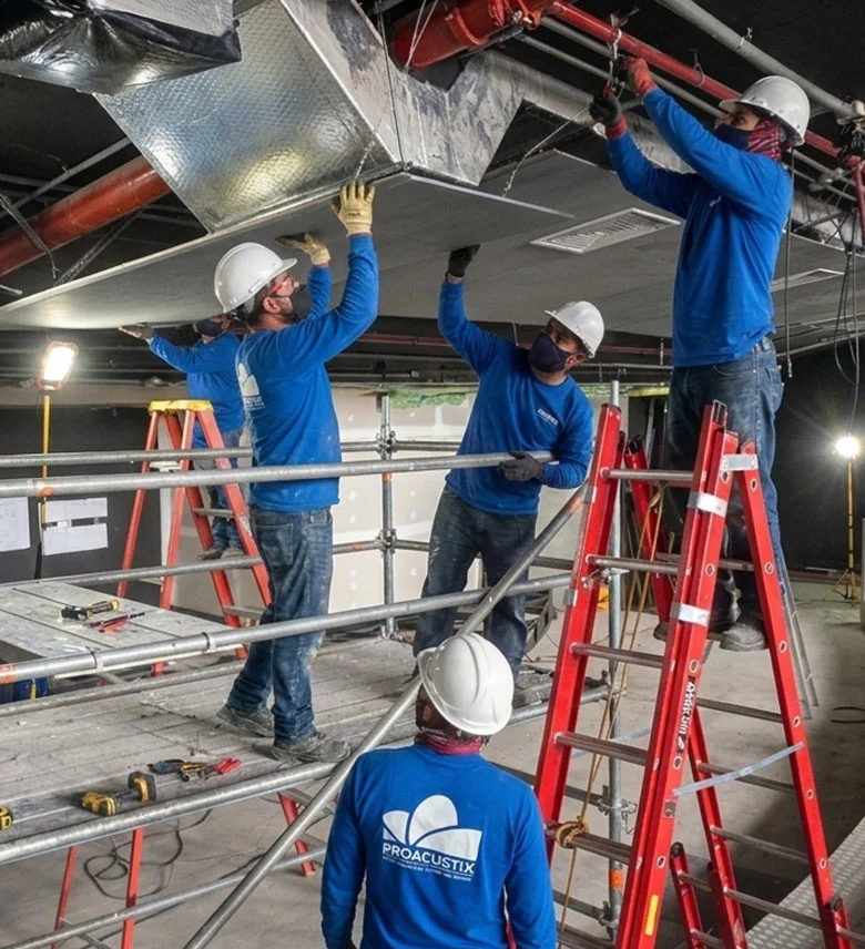Cinco trabajadores con cascos blancos y camisas azules instalando paneles en el techo dentro de una estructura de andamios en una obra civil.
