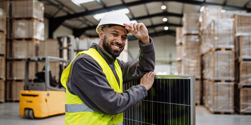 Smiling worker in safety vest and hard hat holding a solar panel in a warehouse with stacked pallets.