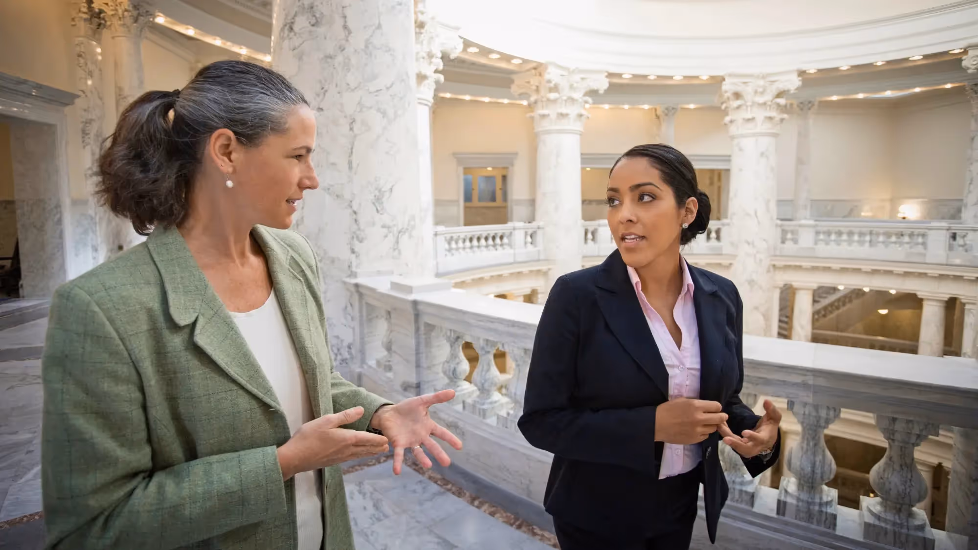 Two professional women in business attire talking inside a marble government building with columns and ornate architecture.