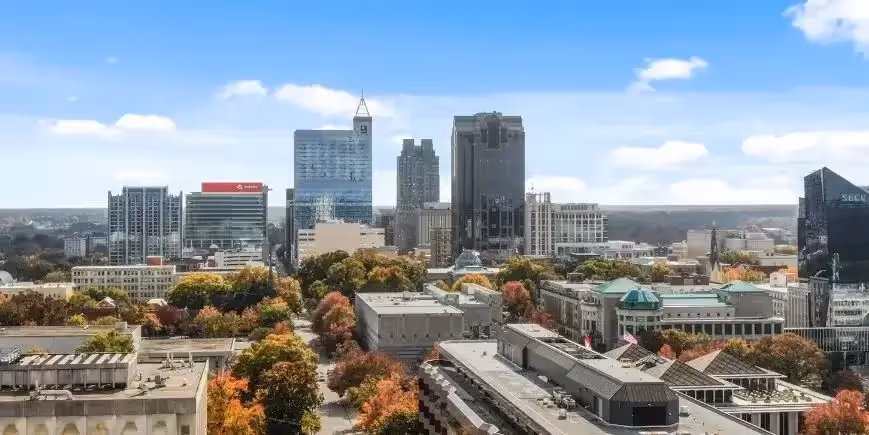Downtown Raleigh skyline with modern high-rises, trees with fall colors, and a clear blue sky with scattered clouds.