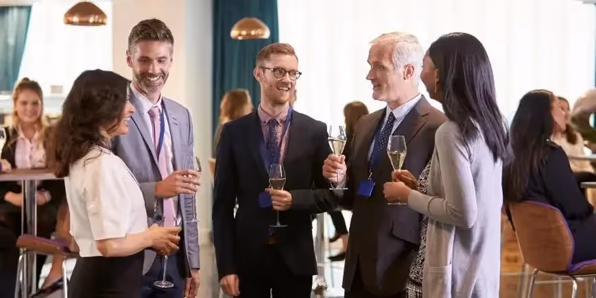 Group of professionals networking at a conference reception, holding drinks and smiling while engaged in conversation.