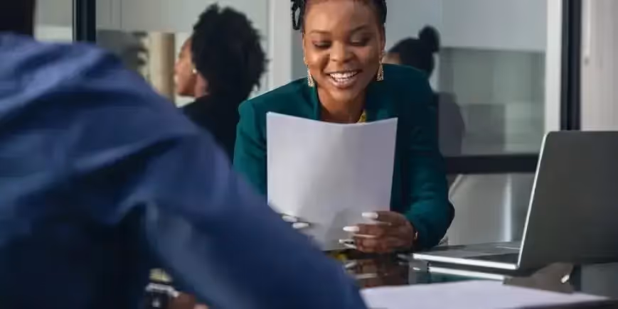 Smiling recruiter reviews a candidate’s resume during an interview at a career fair, with a laptop open on the desk.