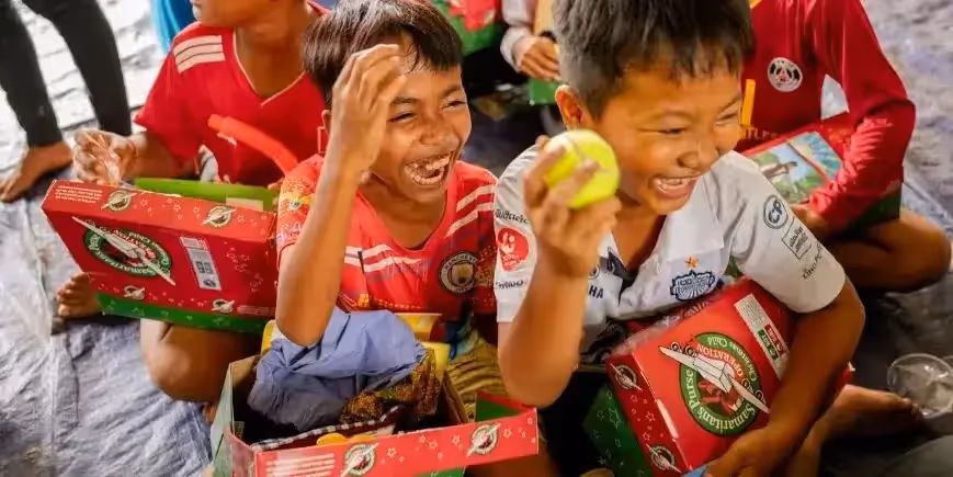 Smiling children joyfully open Operation Christmas Child shoebox gifts, holding toys and items with excitement.
