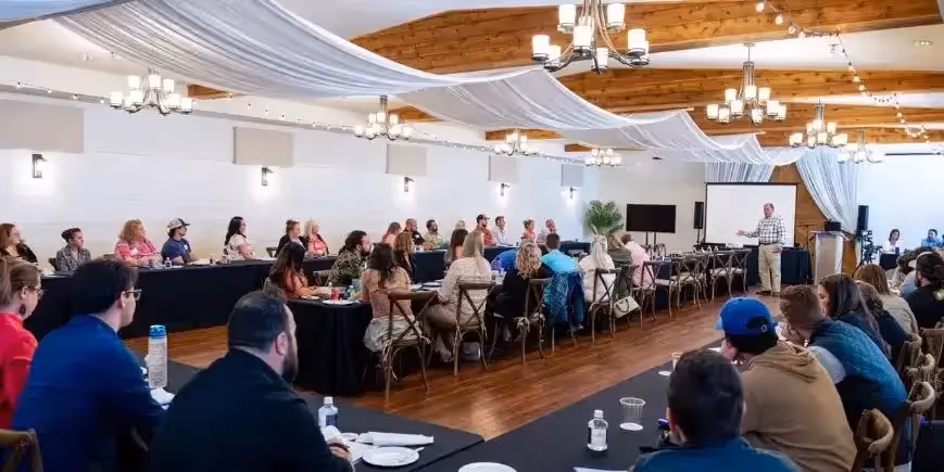 Large group of people seated at tables listening to a speaker presenting at the front of a decorated conference room.