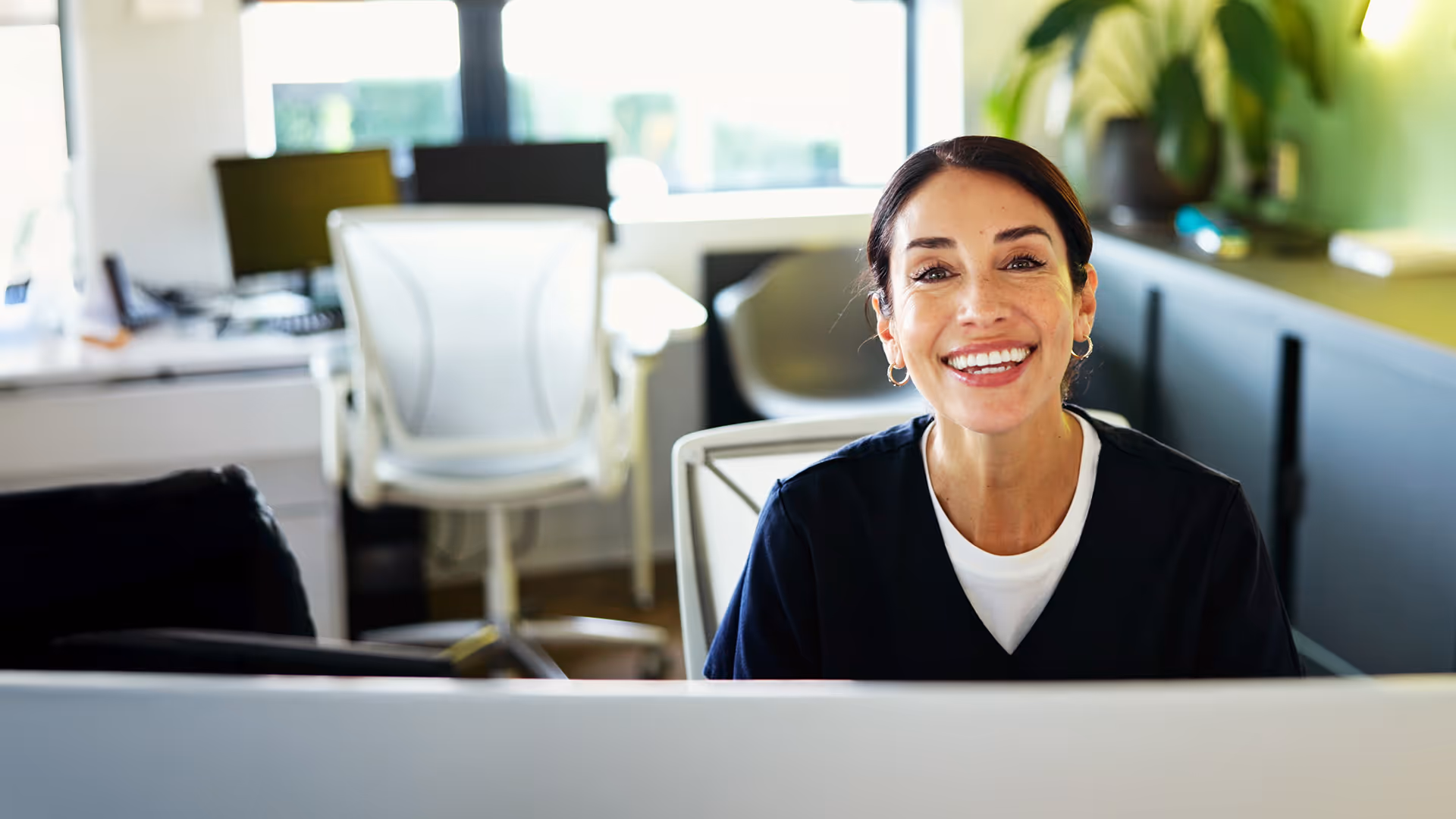 he image shows a cheerful woman sitting at a desk in a bright, modern office. She is smiling warmly, looking directly at the camera, which gives the scene a welcoming and friendly vibe. She has dark hair pulled back, is wearing hoop earrings.