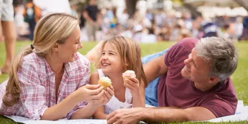 A family enjoys cupcakes together on a blanket in a bustling park.  The image captures a heartwarming moment of a family consisting of a mother, father, and their young daughter. They are lying on a light-blue checkered blanket spread on a lawn.