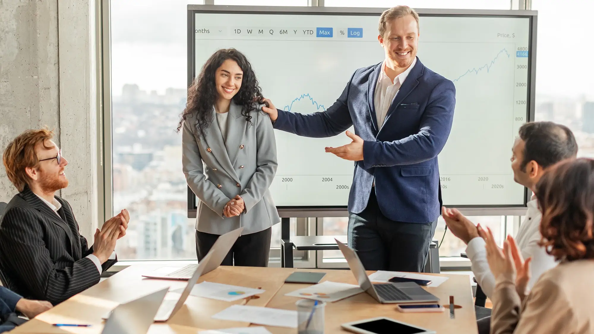 A group of colleagues sit around a conference table applauding as a man standing at the front of the room introduces and praises a smiling woman, with a large screen behind them displaying an upward-trending chart.