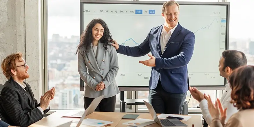 A group of colleagues sit around a conference table applauding as a man standing at the front of the room introduces and praises a smiling woman, with a large screen behind them displaying an upward-trending chart.