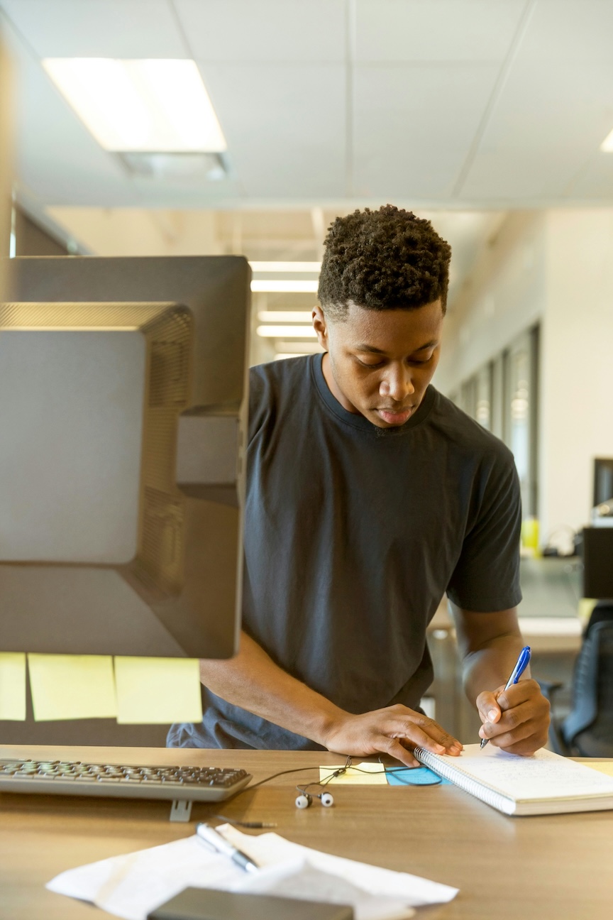 A student writes on a notepad while working at his desk alongside a desktop computer
