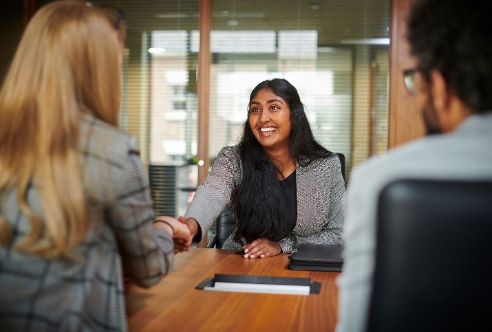 Three professionals sit at a conference table. Two shake hands.