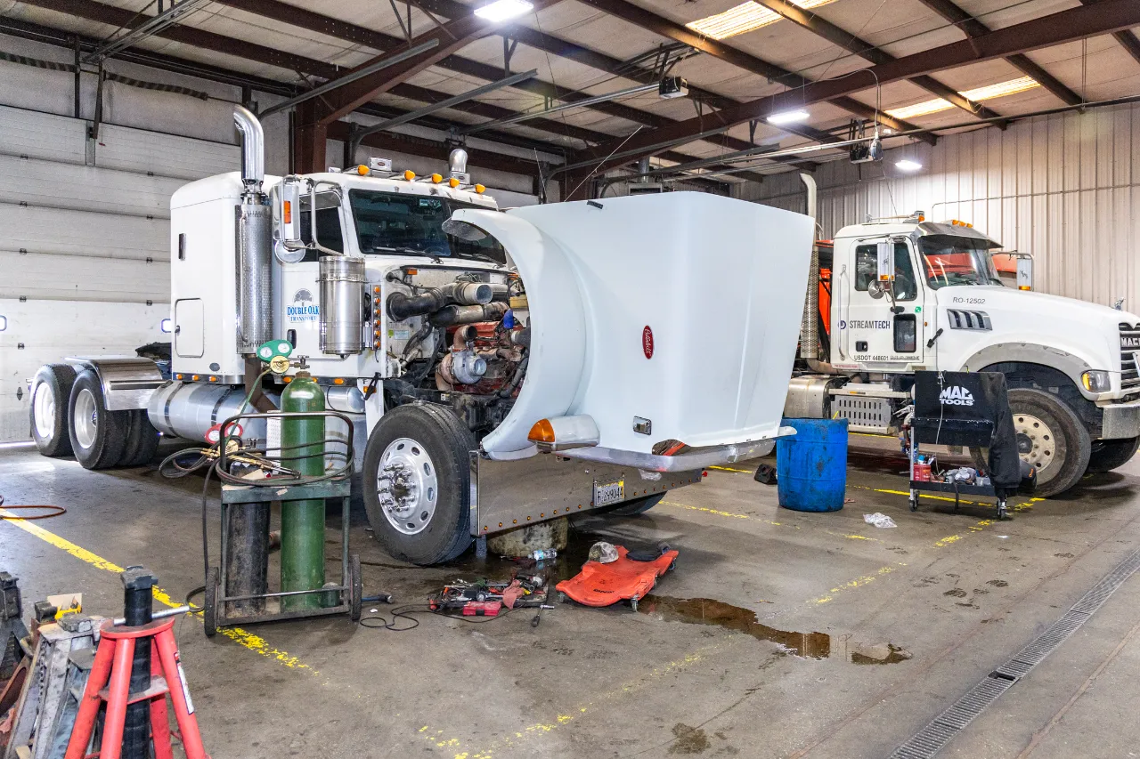 Truck maintenance in progress on a semi with raised hood and exposed engine inside shop; ServiceTech truck visible.