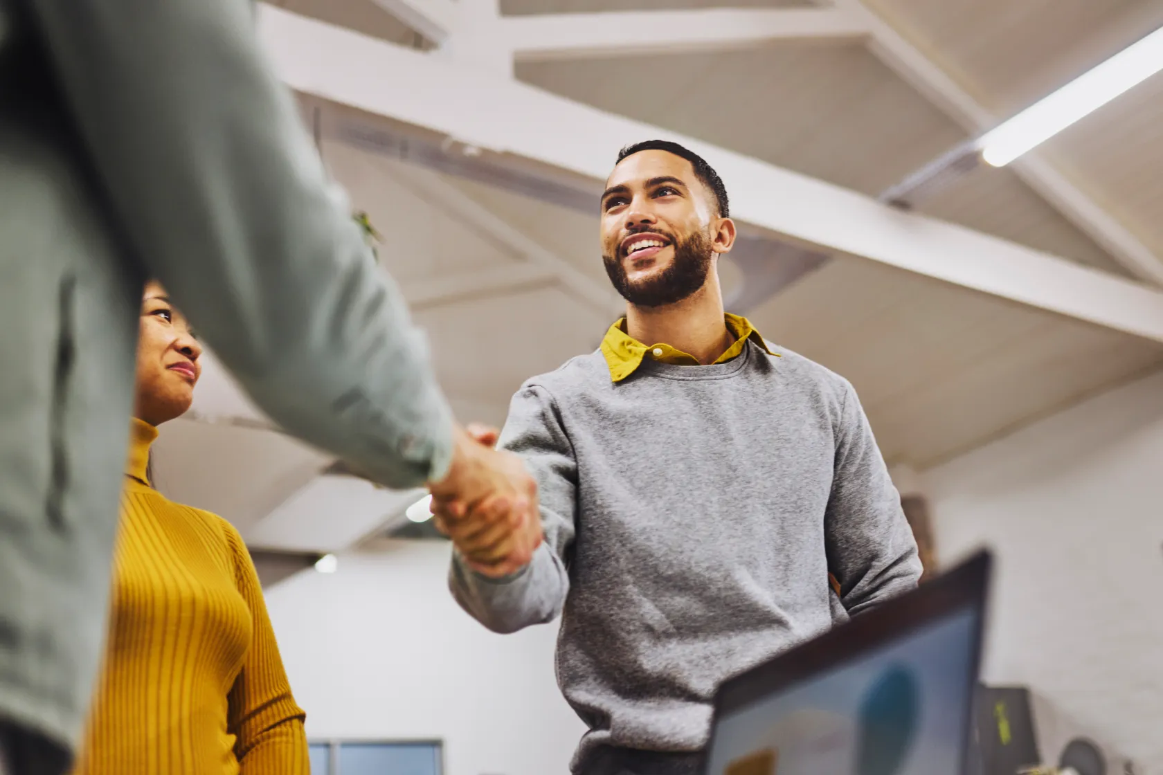 Two colleagues shaking hands in a modern office, with a woman looking on approvingly in the background