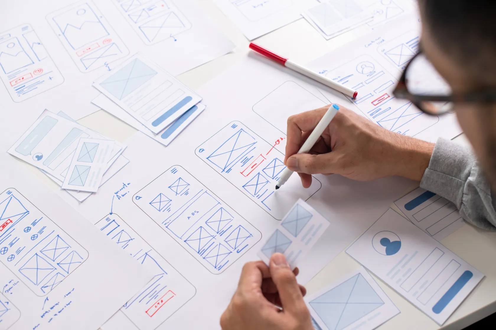 Designer drawing mobile app wireframes and UI layouts spread across a white table
