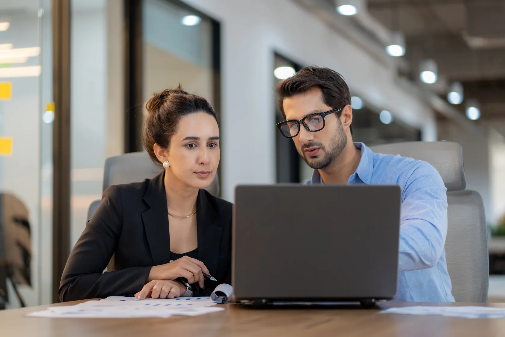 Two colleagues sitting side by side at a desk, focused on reviewing content on a laptop screen
