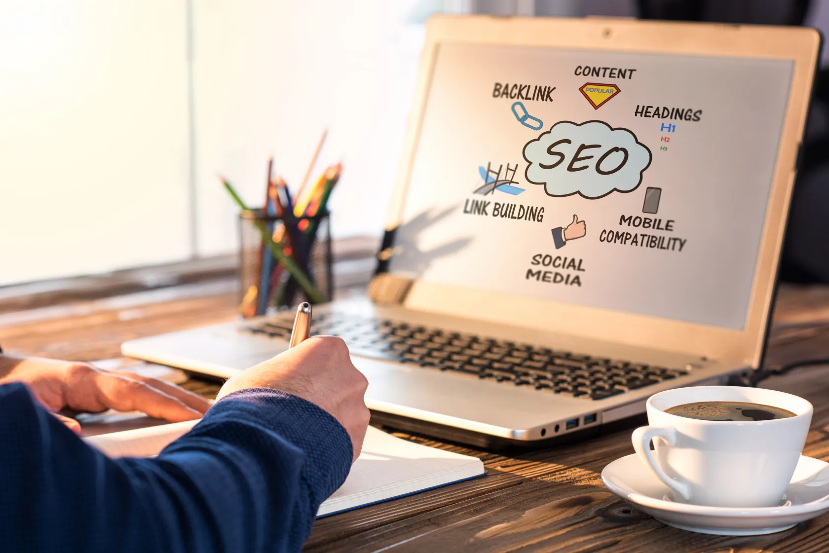 Person taking notes in a notebook while reviewing SEO concepts on a laptop screen, including backlinks, content, headings, link building, social media, and mobile compatibility
