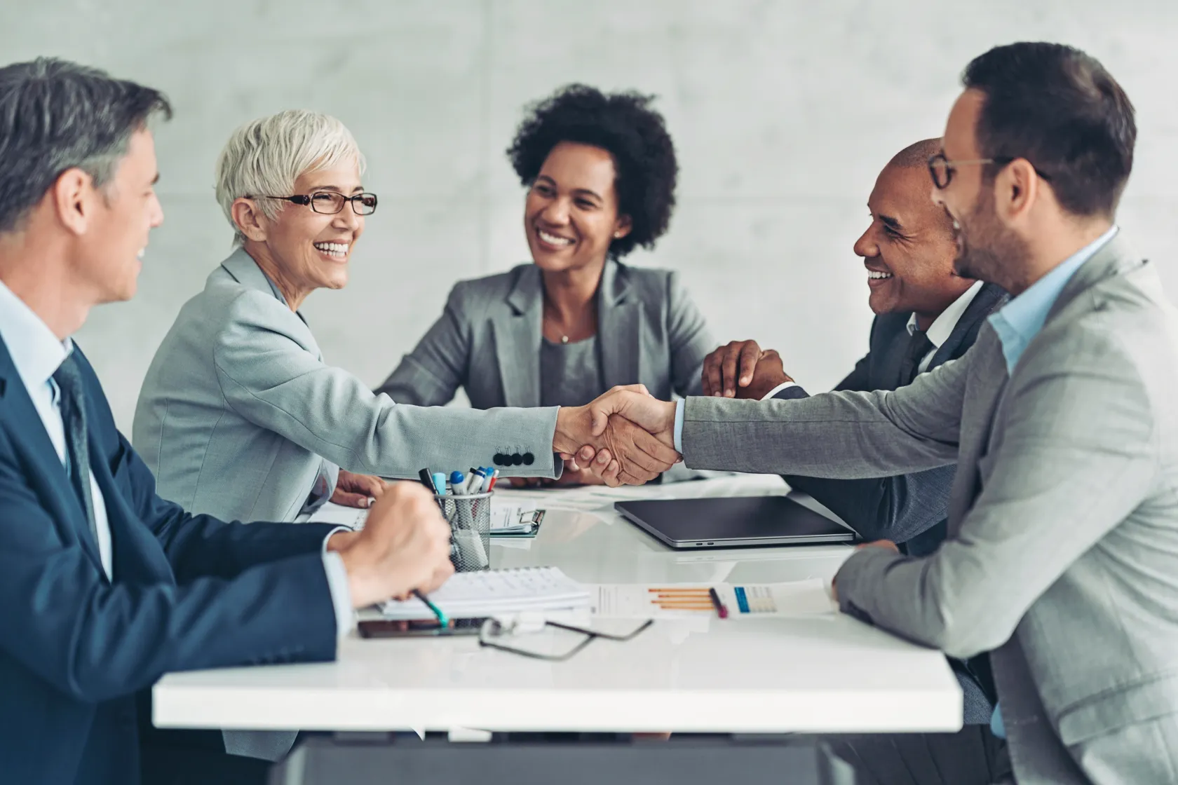 Five diverse business professionals smiling and shaking hands across a conference table, signaling a successful deal