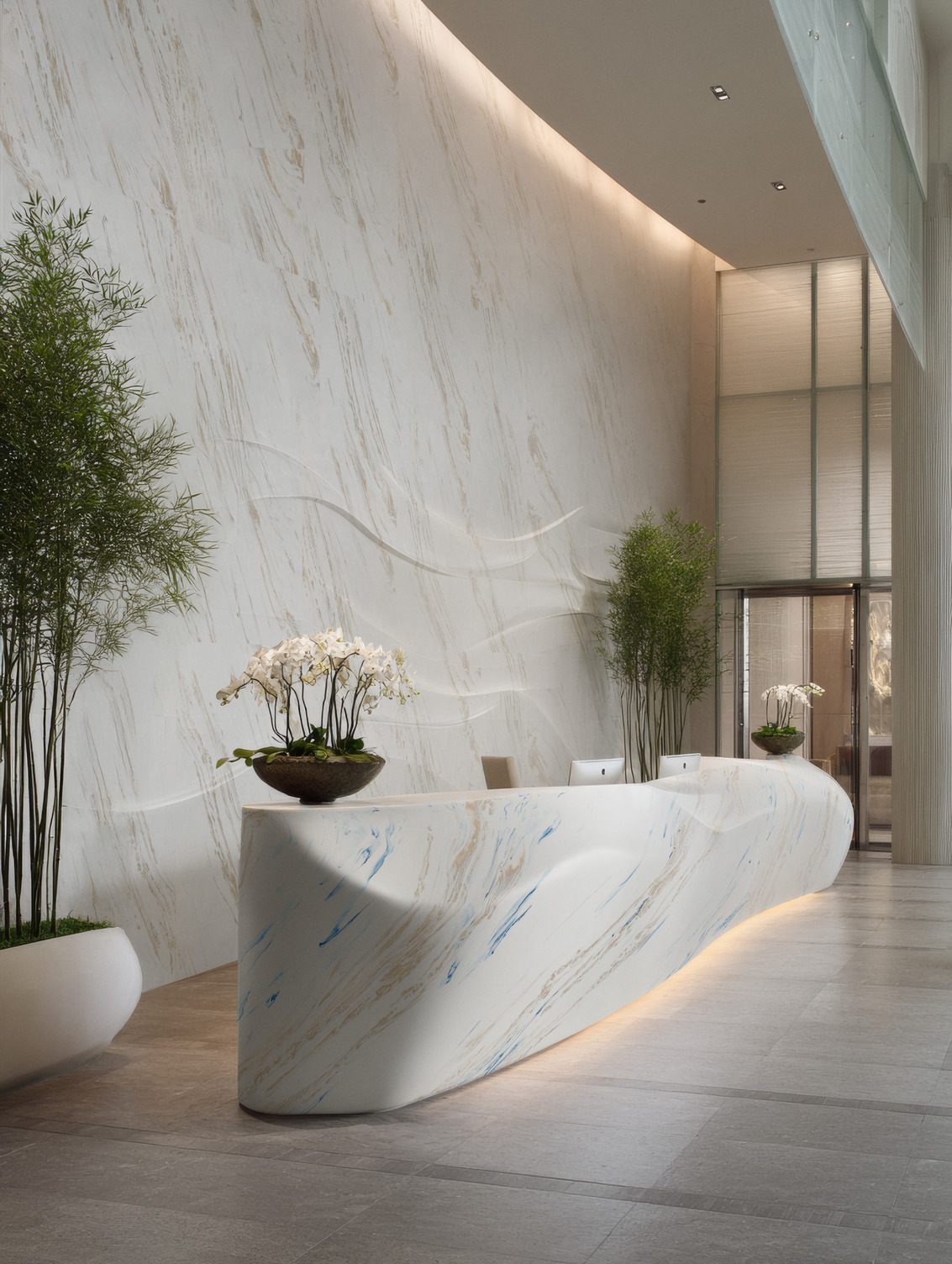 Eleganter Empfangsbereich mit geschwungener, hell marmorierter Rezeption vor einer raumhohen Wand; Pflanzen und Orchideen setzen grüne Akzente. Dekor Wand: Corian® Vasto Greige Dekor Empfangstresen: Corian® Vasto Laguna
