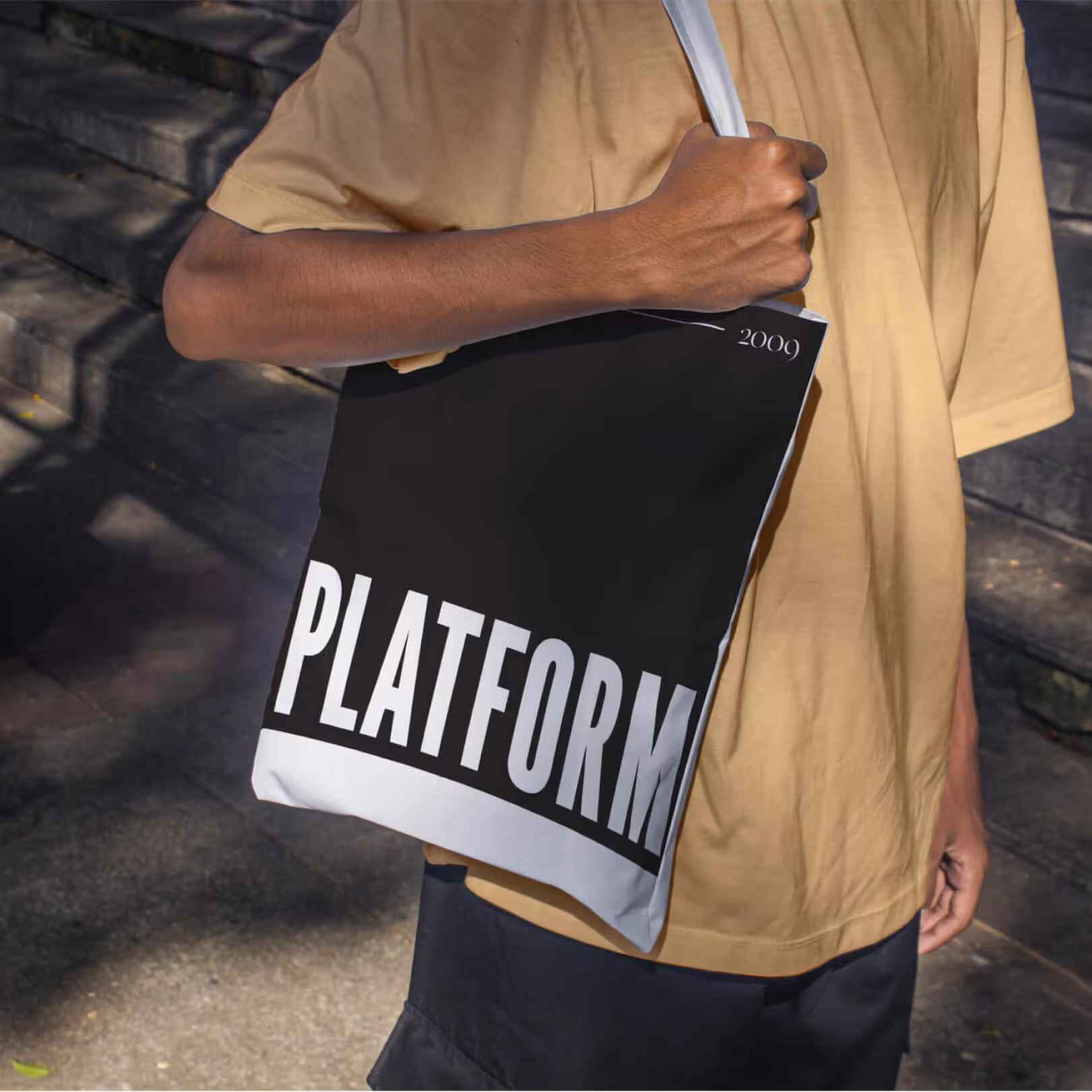 Black and white tote bag printed with the Platform Studios logo, carried by a person wearing a tan shirt outdoors.