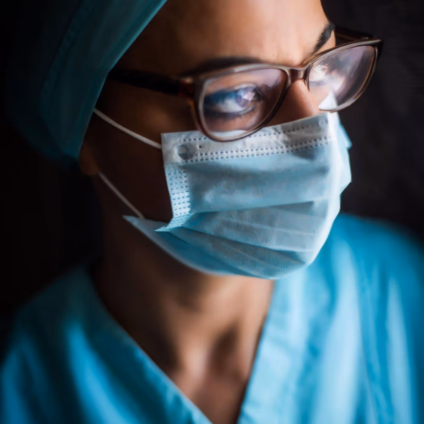 Female surgeon in teal scrubs and glasses wearing surgical mask in focused concentration