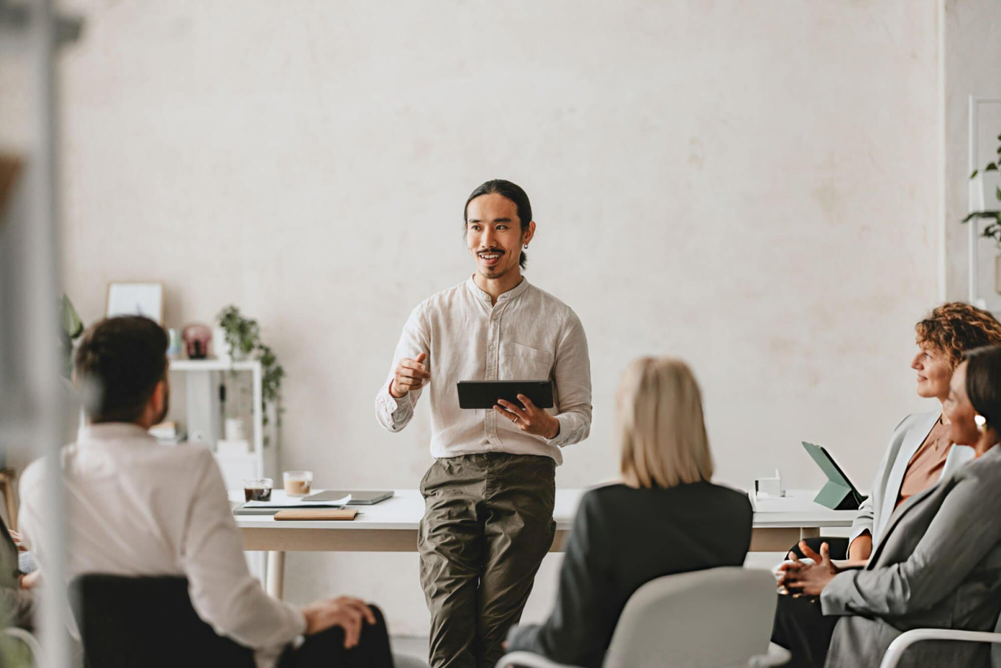 Man holding a tablet speaking to a group of four seated people in a modern office setting.