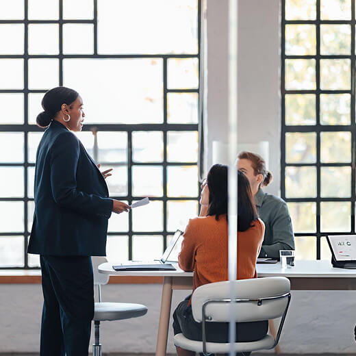 Businesswoman standing and speaking to two seated colleagues in a modern office with large windows.