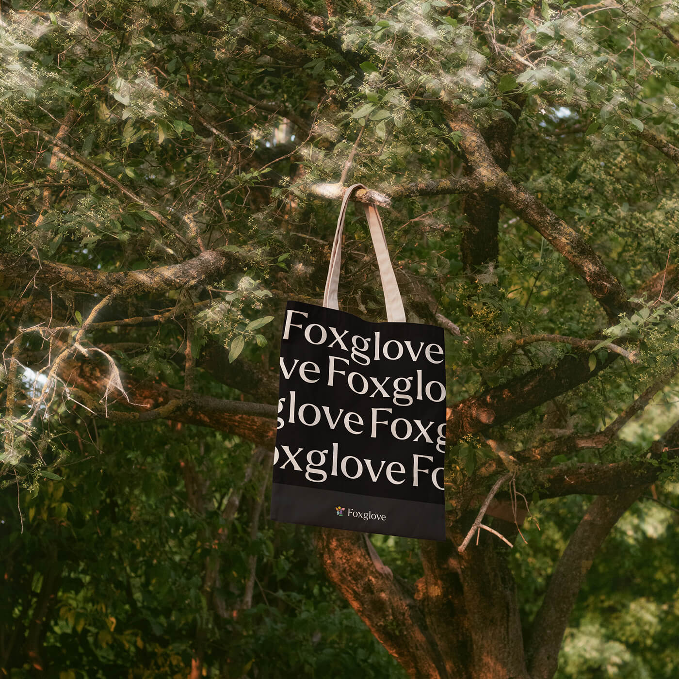 Black tote bag with repeated white 'Foxglove' text and a small multicolored logo hanging from a tree branch in a green forest.