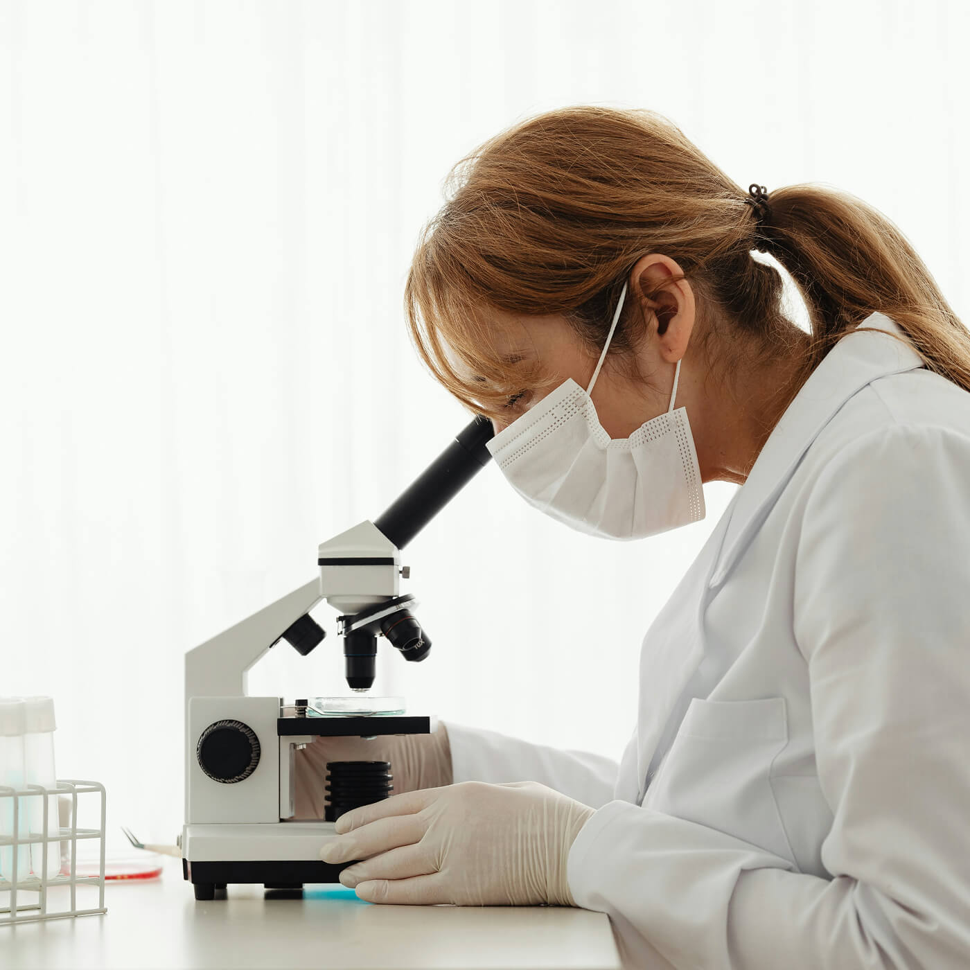 Scientist wearing a face mask and gloves looking through a microscope in a laboratory.