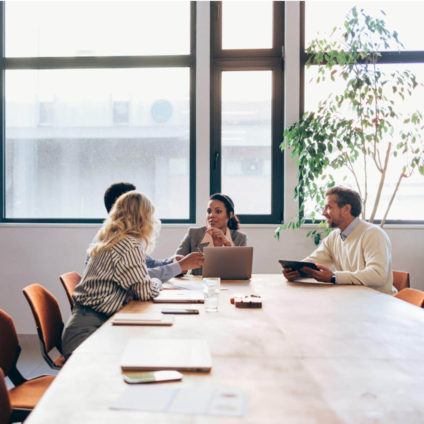 Four professionals engaged in a meeting around a conference table with laptops and documents in a bright office with large windows.