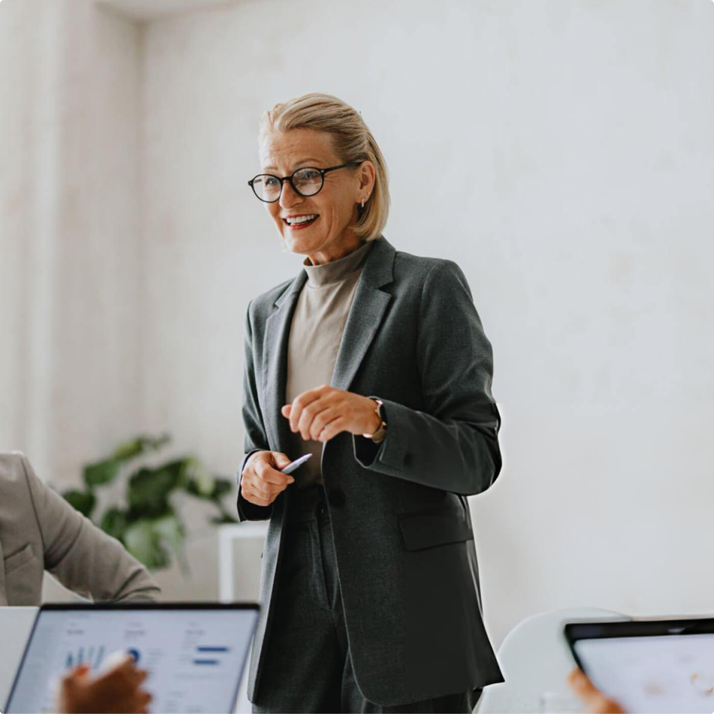 Smiling older woman in glasses and gray suit speaking in a modern office with colleagues using laptops.