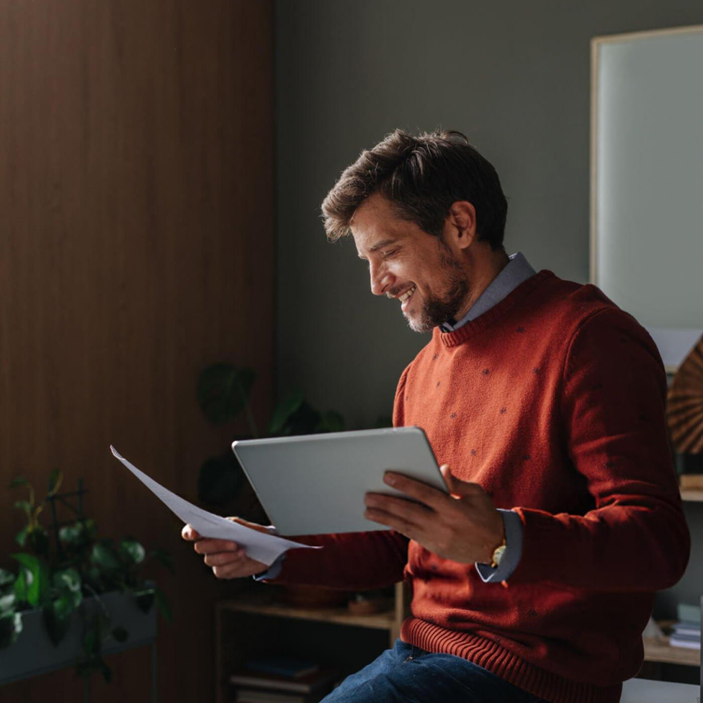 Smiling man in an orange sweater holding a tablet in one hand and a paper in the other while sitting indoors.
