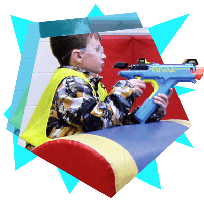 Child wearing safety goggles and a yellow cape aiming a blue Nerf Rival toy gun indoors.