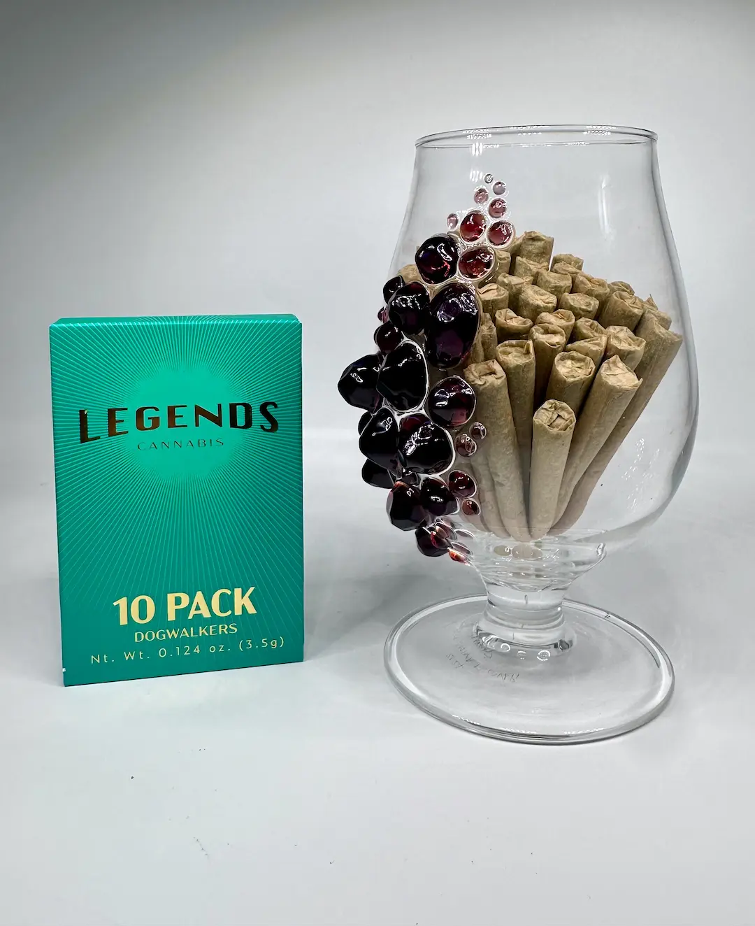 Legends 10 pack cannabis dogwalkers package next to a glass cup with a bunch of pre-rolls inside the glass all on a light gray background