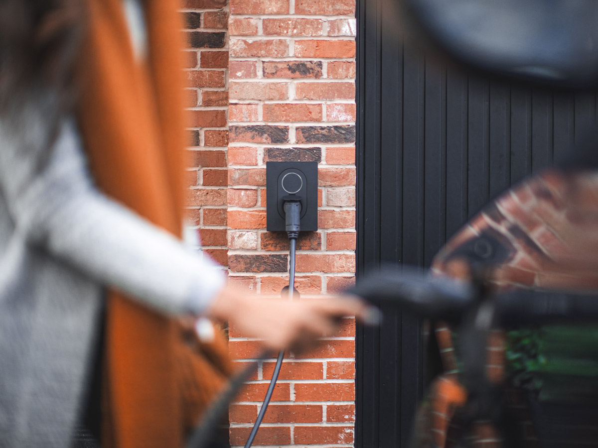 Image of a blurred female in the foreground plugging in an electric car, in the background you can see the wall unit for the charger
