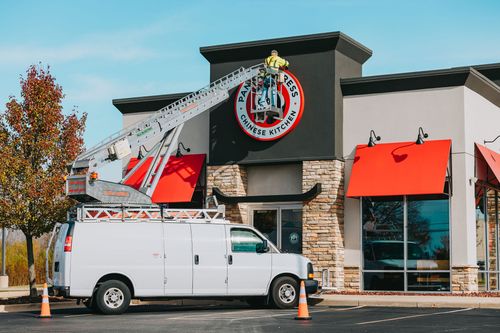 Worker in a yellow jacket installing or repairing a Panda Express Chinese Kitchen sign on a building using a white van with a ladder lift.
