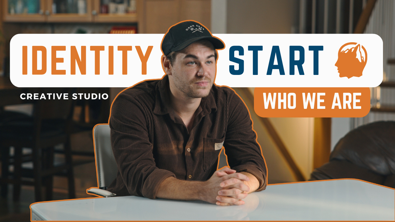 Young man wearing a dark shirt and black cap sitting at a white table in a creative studio setting with text reading 'Identity Start Who We Are'.