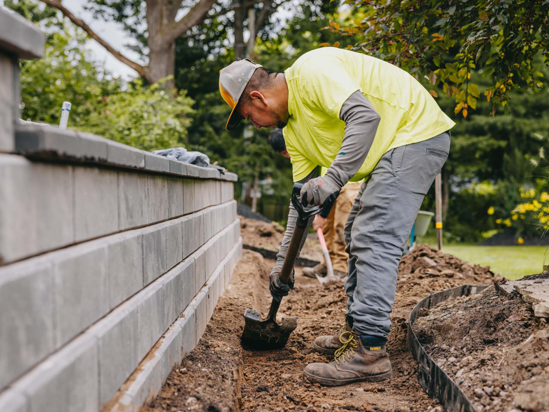 Landscaper digging soil foundation beside retaining wall during landscape installation