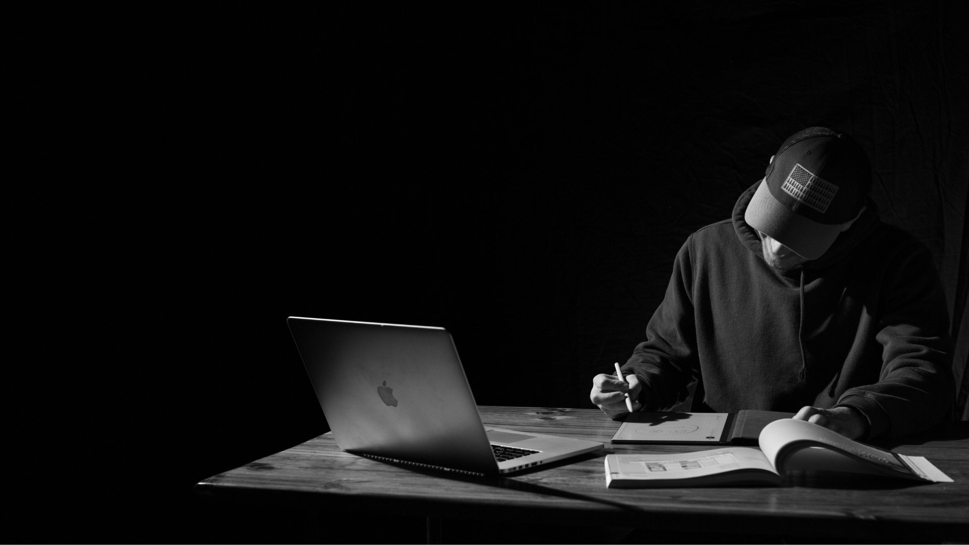 Person in a cap working on a laptop and tablet at a wooden table in low light, planning a website.
