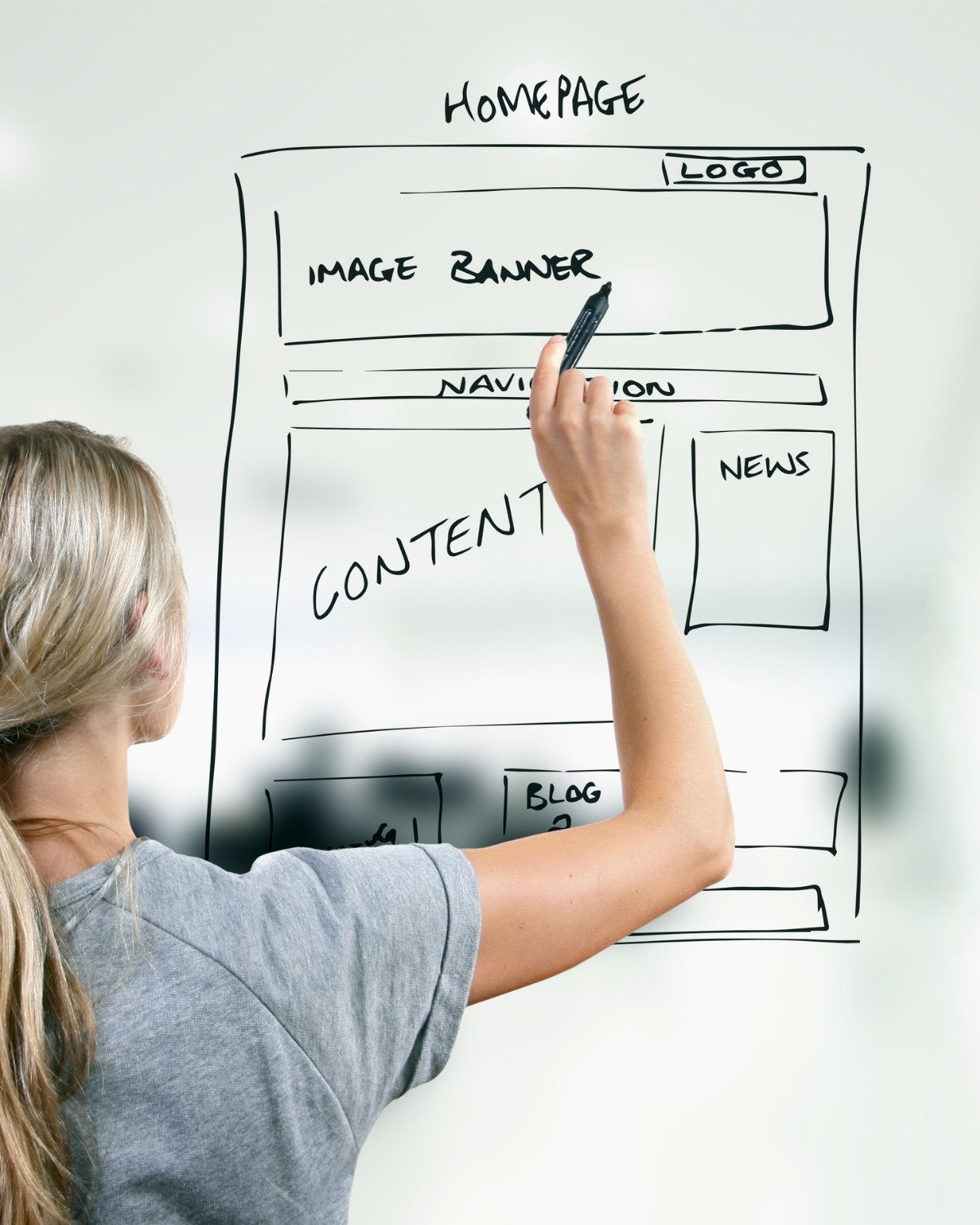 Person sketching a website homepage layout on a transparent board with logo, banner, navigation and content sections.