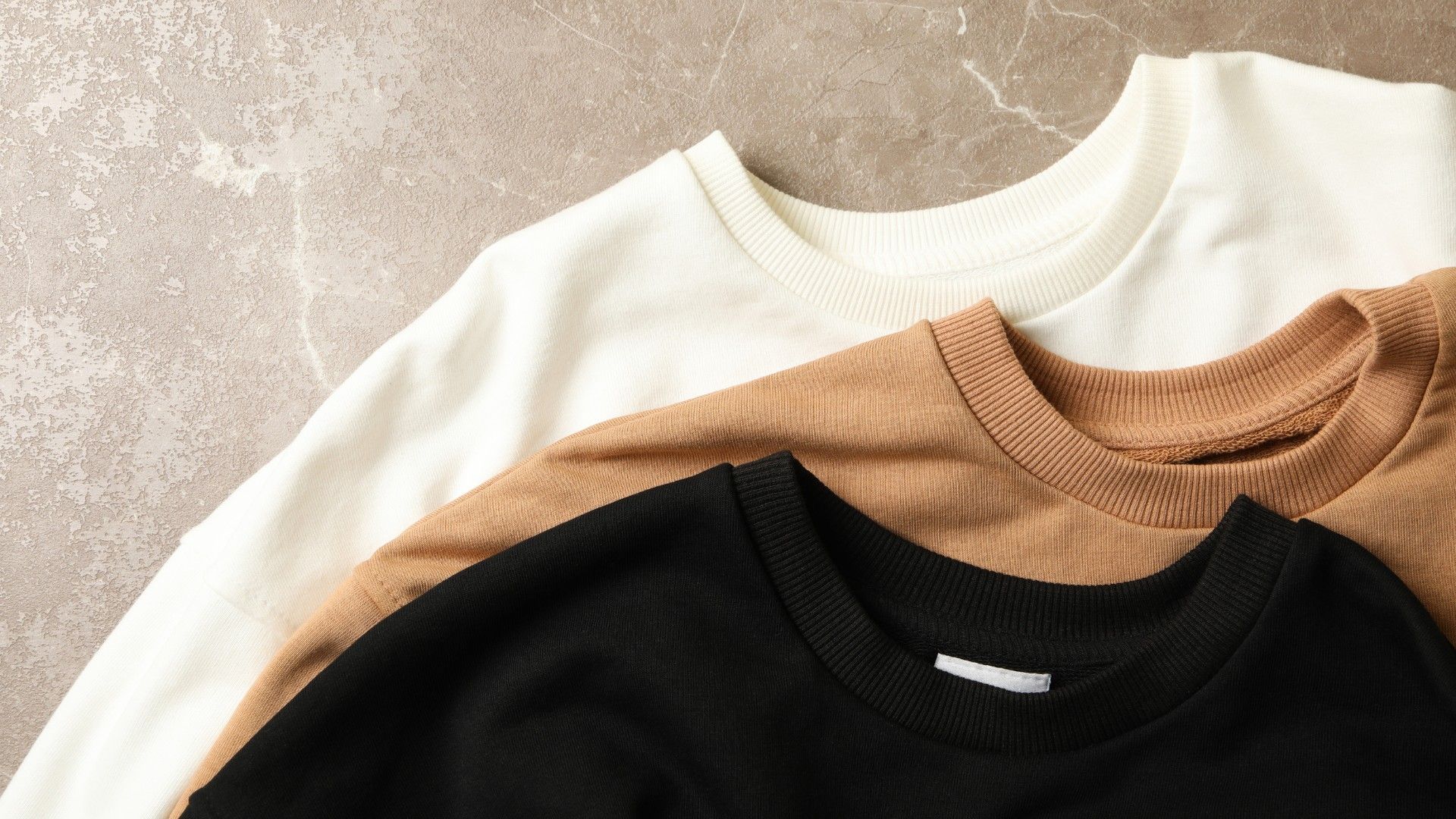 Folded cream, tan and black crewneck sweatshirts stacked on a stone surface